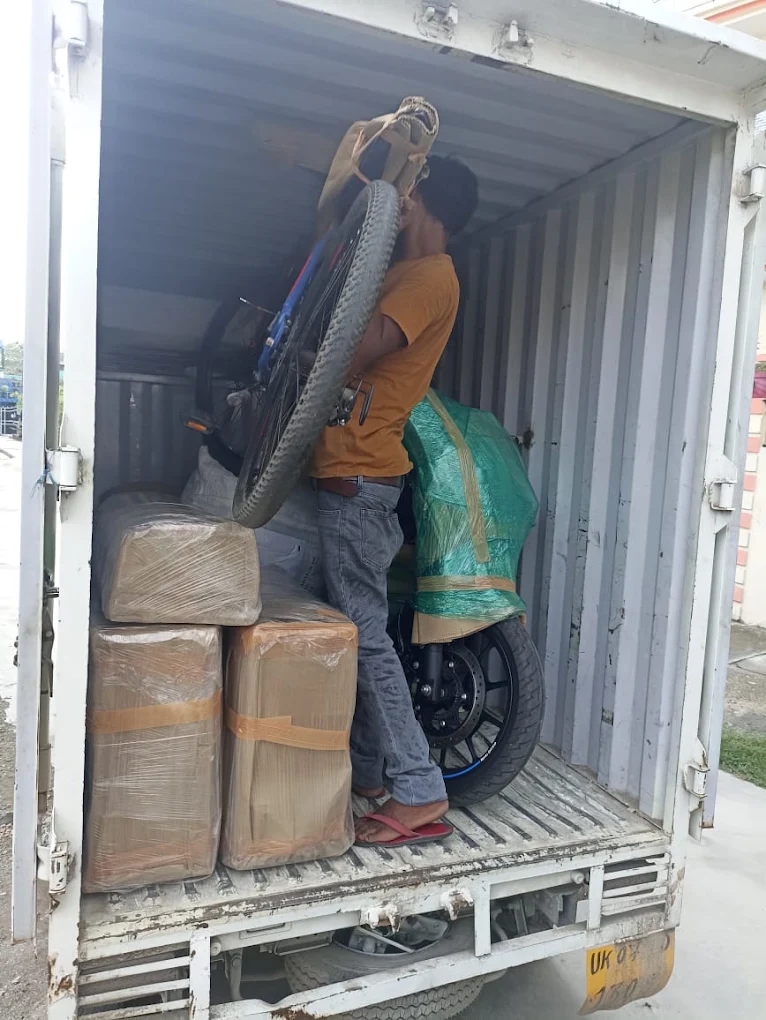 A worker loading packed household goods and a wrapped two-wheeler into a moving truck
